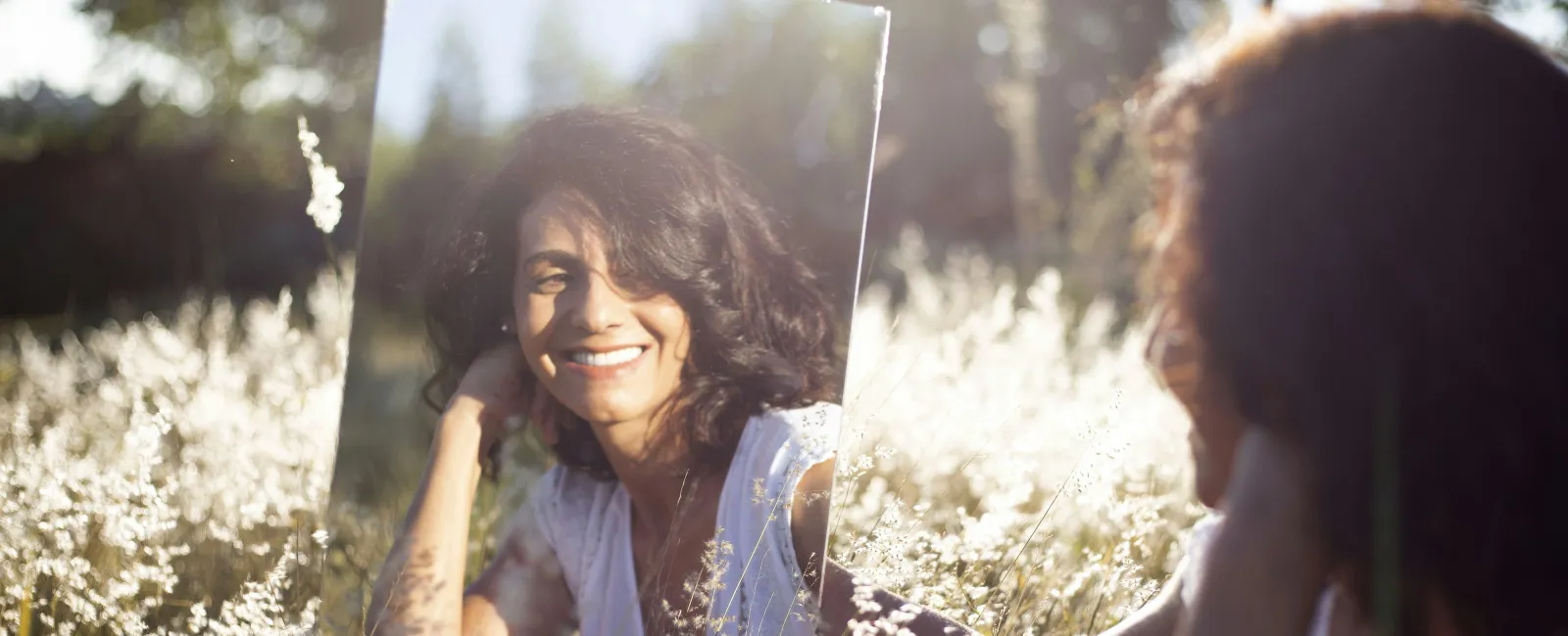 Woman with curly hair smiling at her reflection in a mirror outdoors in sunlit wildflowers.