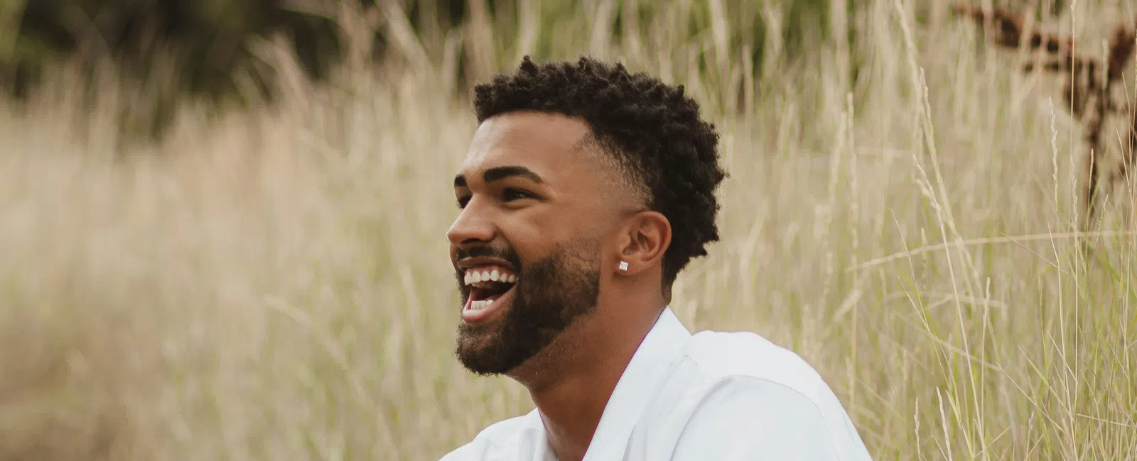 Smiling man in white shirt and khaki shorts sitting outdoors in tall grass looking happy and relaxed.