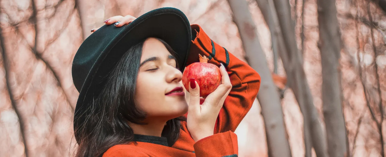 Woman in black hat and orange sweater holding a red apple in a forest with autumn leaves and bare trees.