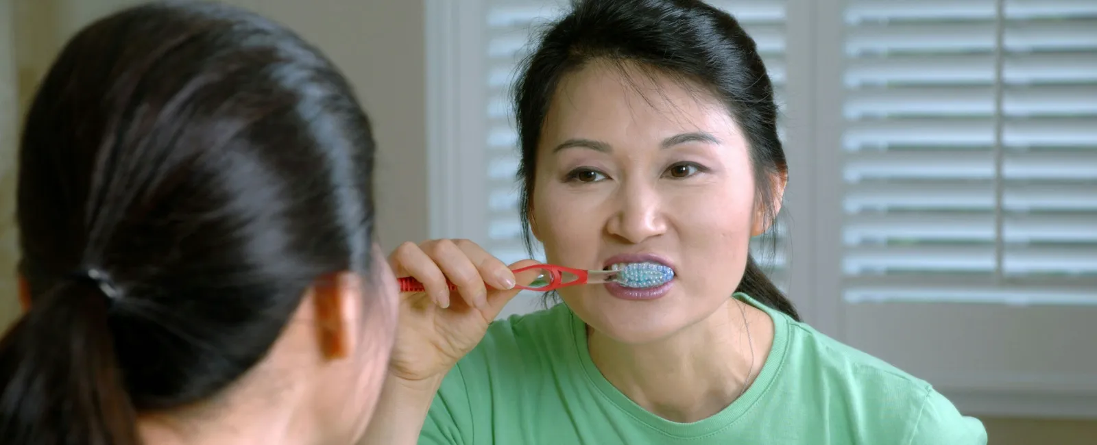 Woman in green shirt brushing her teeth looking into the bathroom mirror with white blinds in background