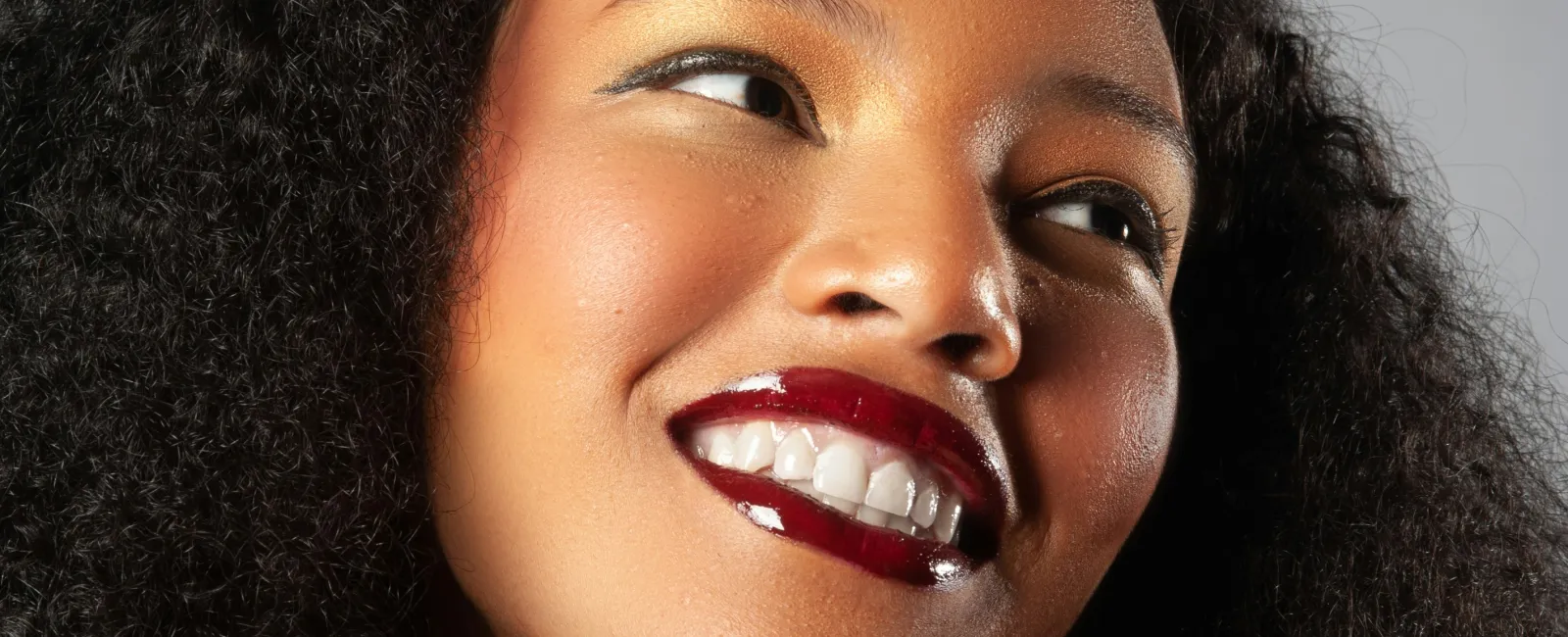 Close-up of a smiling woman with curly hair, glossy dark red lipstick, and golden eye makeup against a gray background.