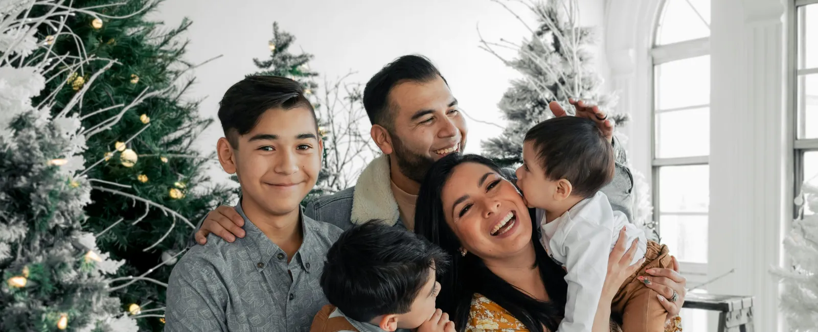 Smiling family of five hugging and enjoying together in a bright room decorated with Christmas trees and lights.