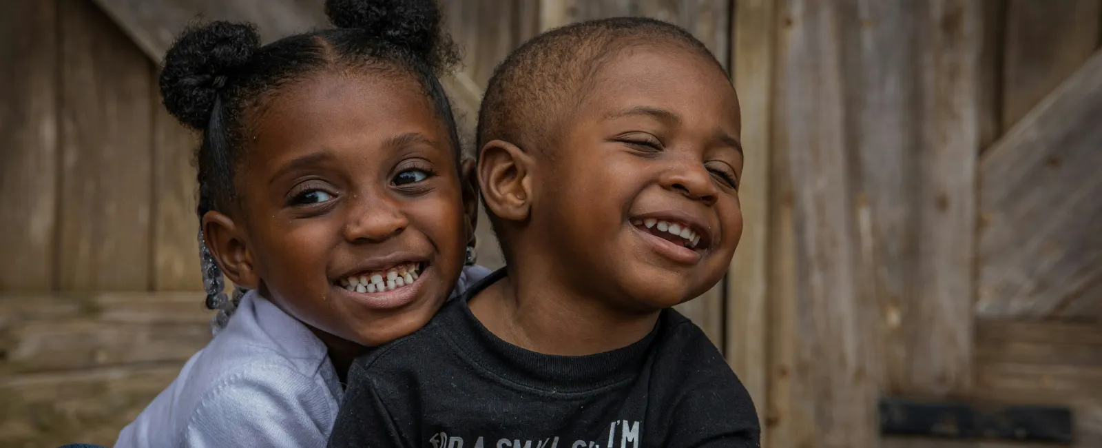 Two happy children smiling and hugging in front of a rustic wooden backdrop outdoors.