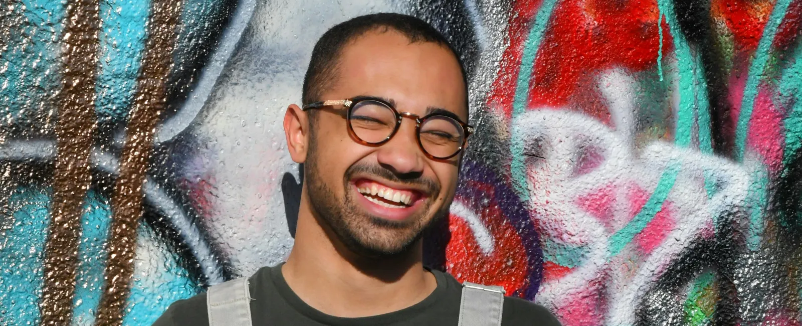 Smiling man wearing glasses and gray overalls stands against a colorful graffiti wall holding a black cap.