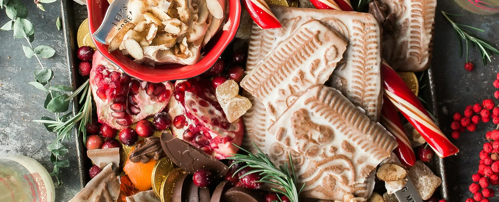 Holiday dessert tray with chocolate, gingerbread, candy canes, pomegranate, fruit, and festive herbs on rustic table.