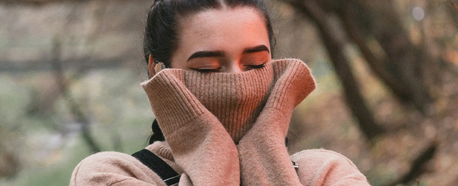 Woman in a cozy beige sweater covering her face with sleeves in a blurred autumnal forest setting.