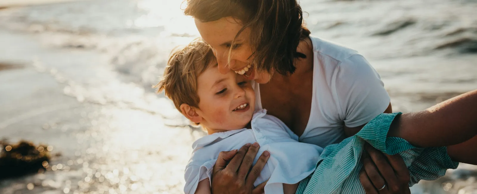 Mother joyfully holding her smiling son at beach during sunset with ocean waves in background