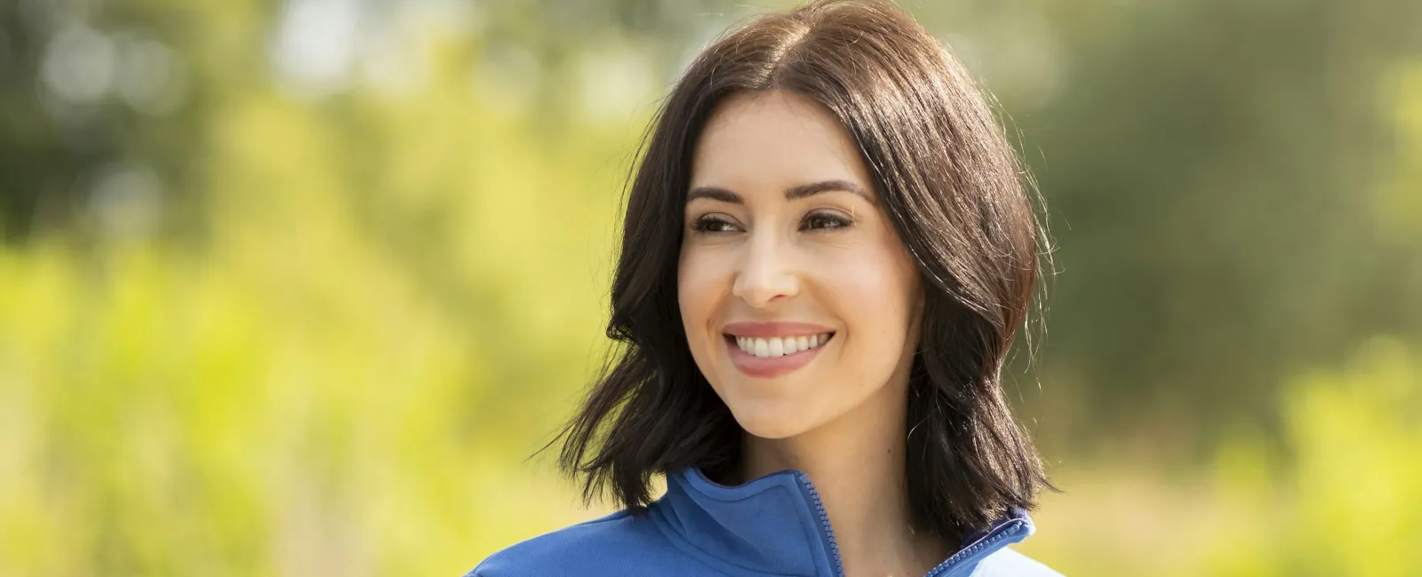 Smiling woman wearing a blue pullover with white and navy stripes, standing outdoors near water and greenery.
