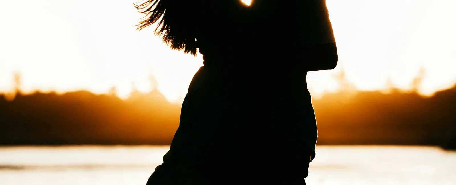 Silhouette of a couple embracing by the water during a golden sunset with flowing dress and wind in hair.