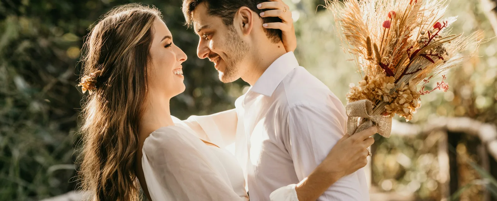 Couple embracing outdoors on a sunny day with a dried floral bouquet in hand, smiling lovingly at each other.
