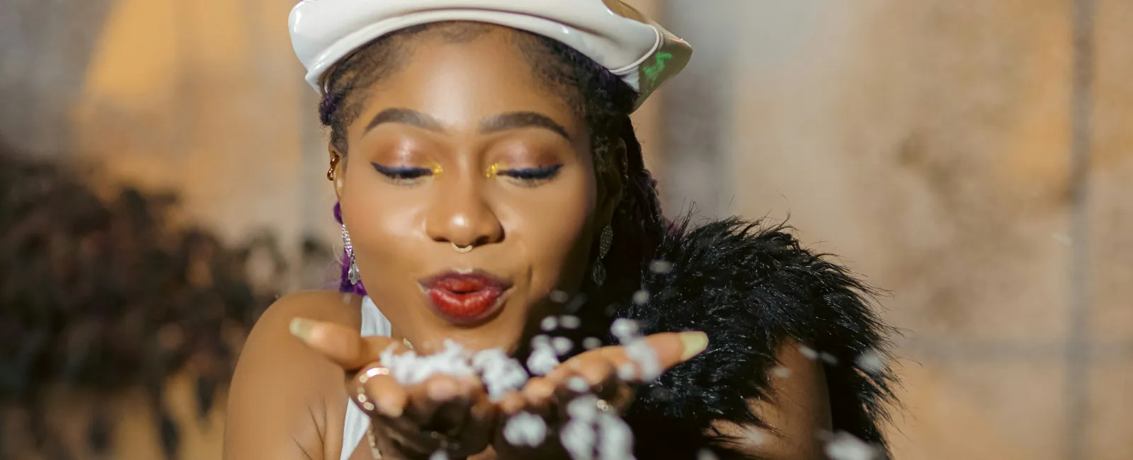 Young woman in white dress and beret blowing confetti from hands with blurred lights in background.