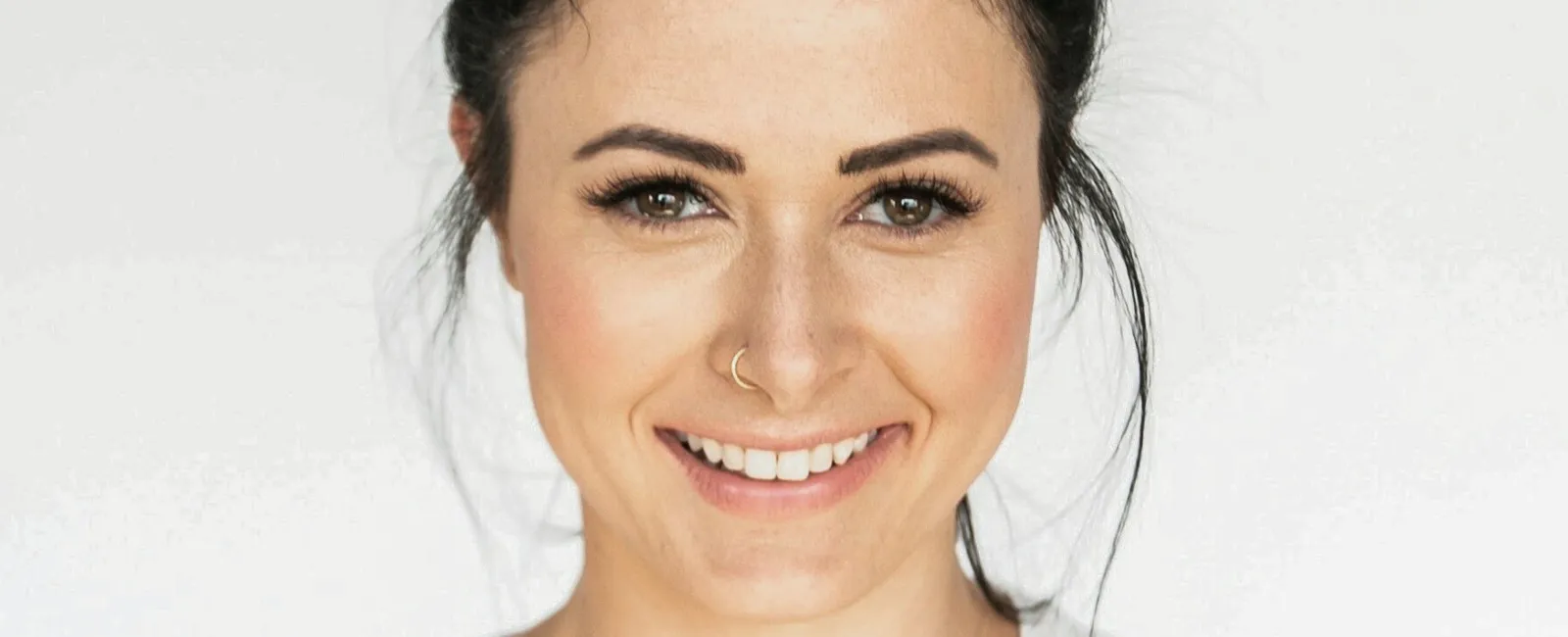 Smiling young woman with dark hair tied in a messy bun wearing a white shirt against plain background