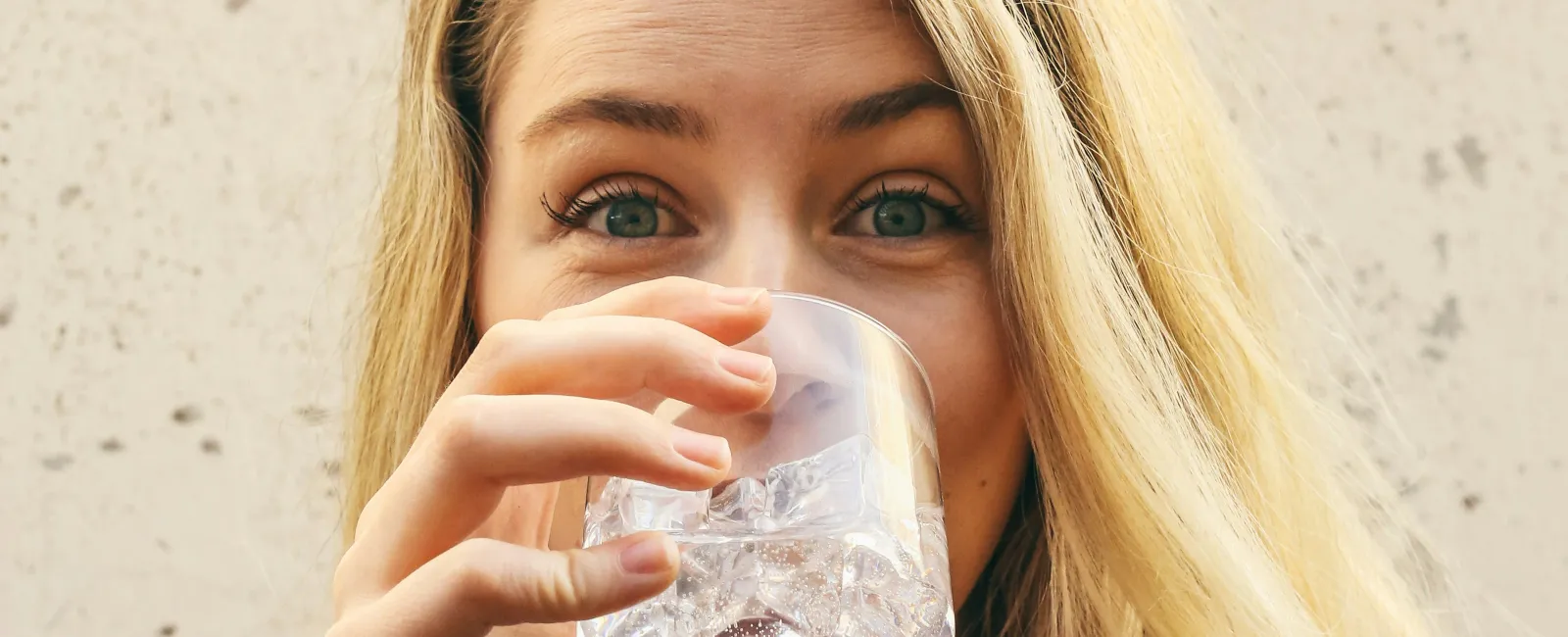 Blonde woman with long hair drinking a glass of water with ice cubes against a beige background