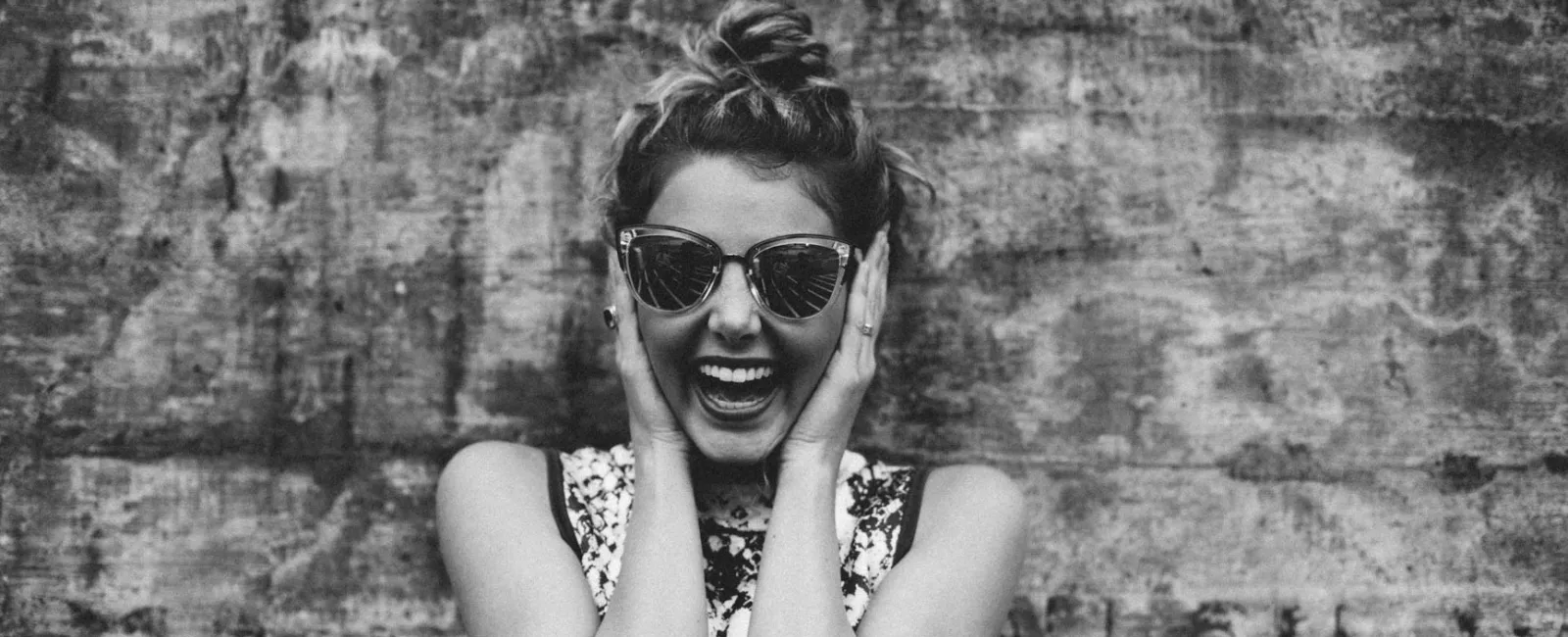 Happy woman wearing sunglasses and a patterned dress smiling against a textured wall background in black and white