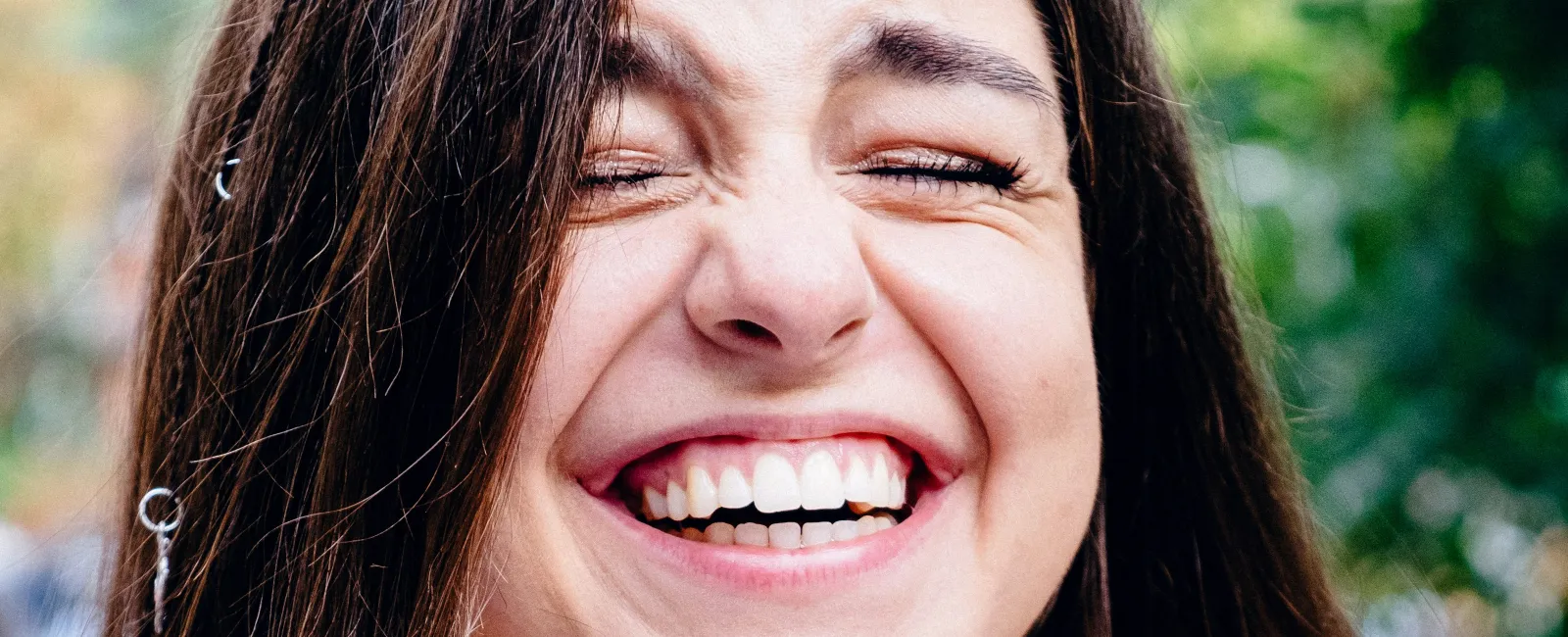 Young woman with long brown hair laughing joyfully outdoors in a sleeveless black top with blurred background.