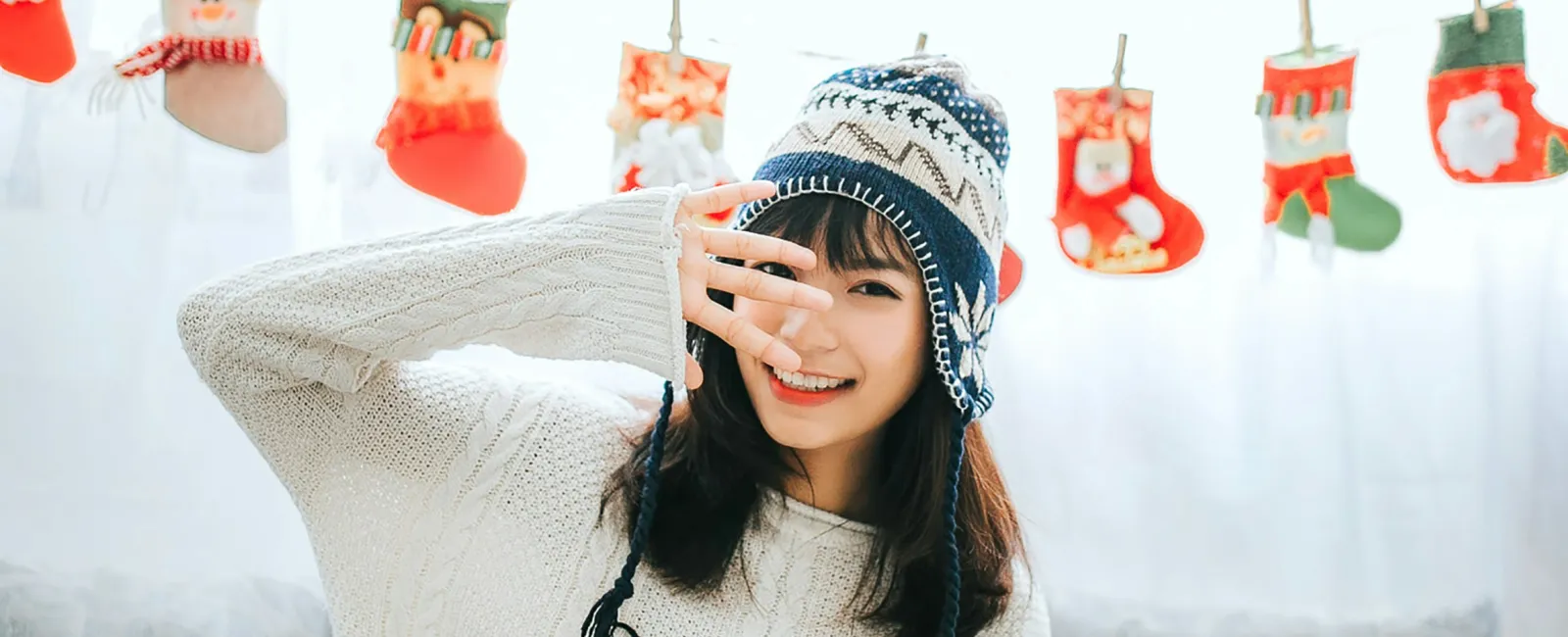 Young woman in cozy sweater and winter hat sitting on couch with festive Christmas stockings behind her.
