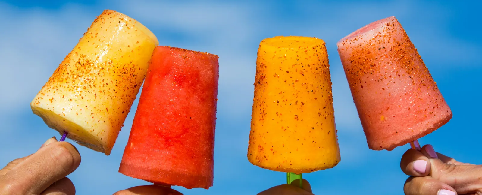 Four hands holding colorful fruit popsicles sprinkled with chili powder against a clear blue sky.