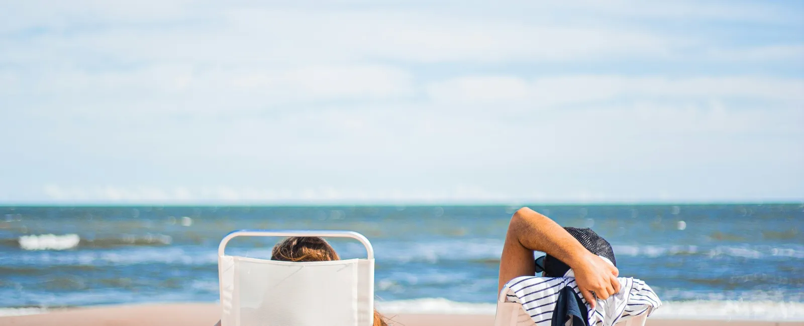 Two people relaxing on white beach chairs facing the ocean under a clear blue sky on sandy shore.