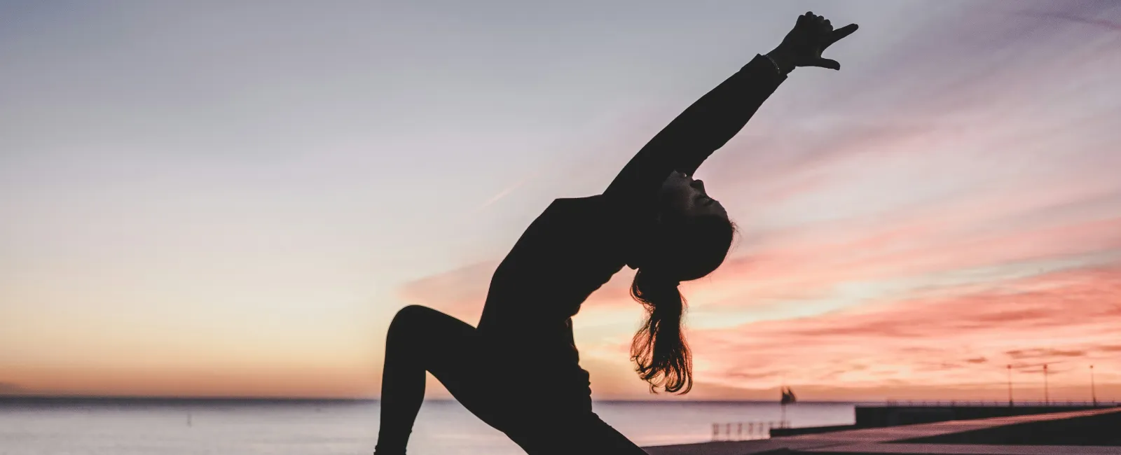 Silhouette of a woman practicing yoga on a mat at sunset by the water with a colorful sky.