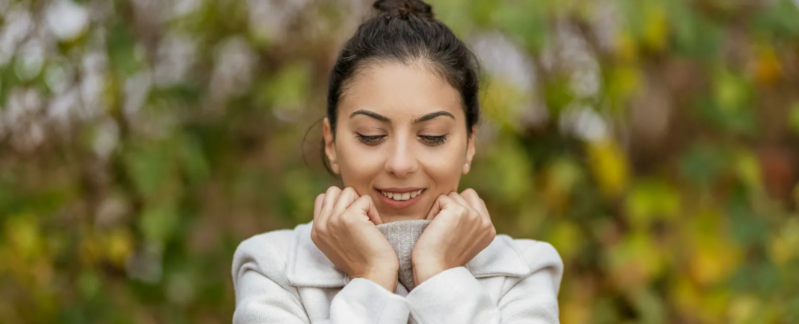 Smiling woman wearing a white coat outdoors with autumn leaves blurred in the background