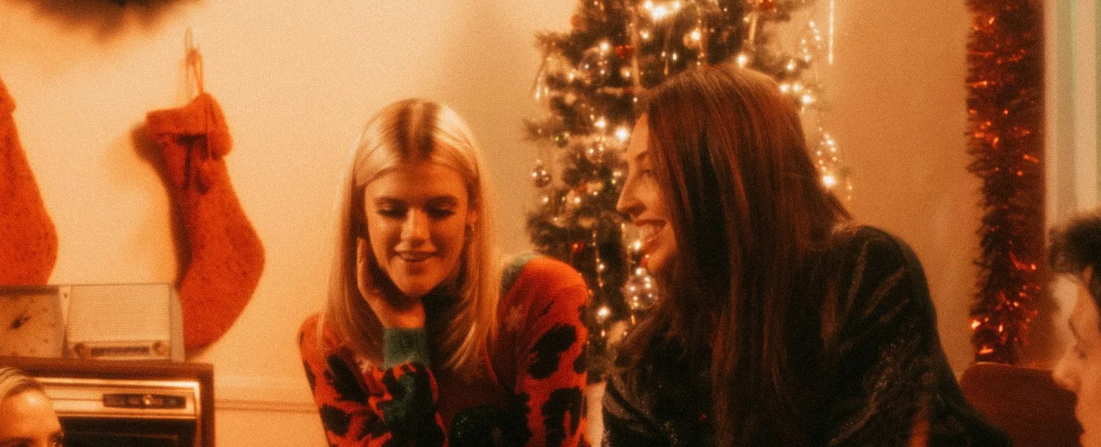 Friends exchange Christmas gifts near decorated tree with stockings and candlelight in cozy holiday setting.