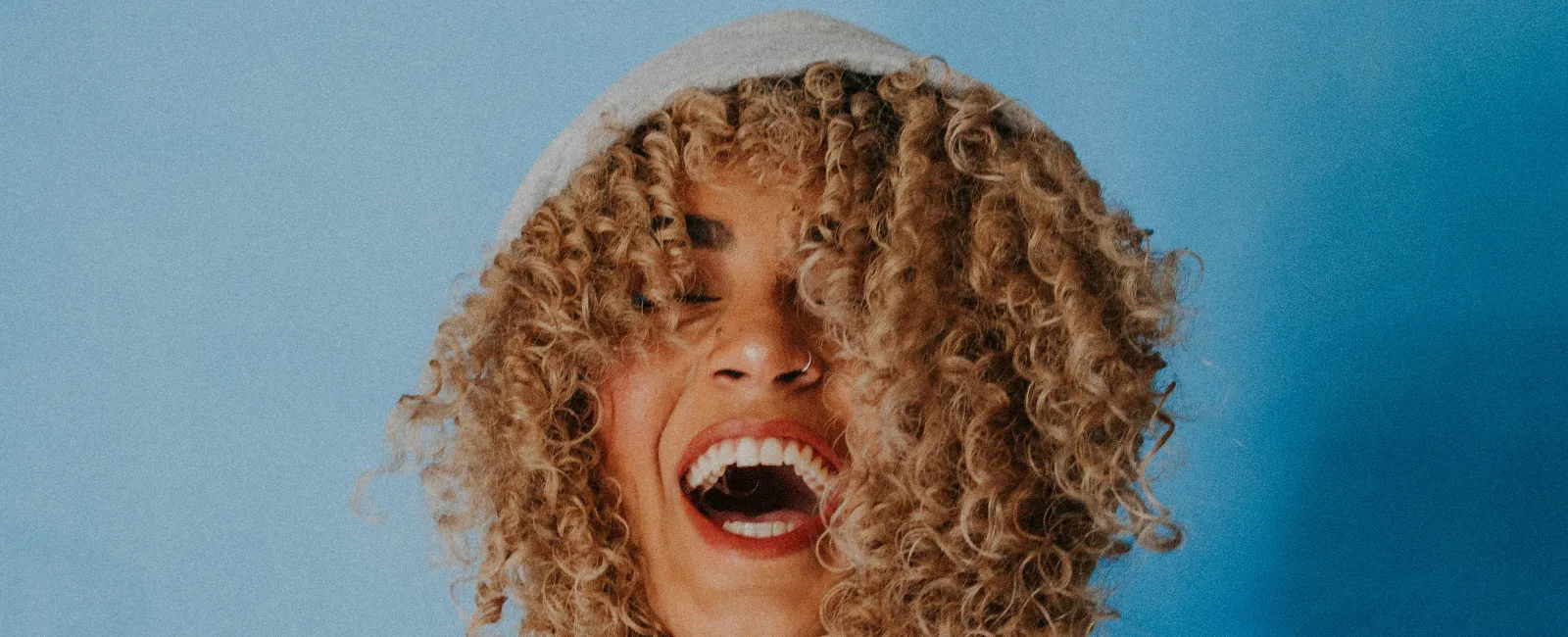 Joyful woman with curly hair laughing while holding the hood of her gray sweatshirt against a blue background