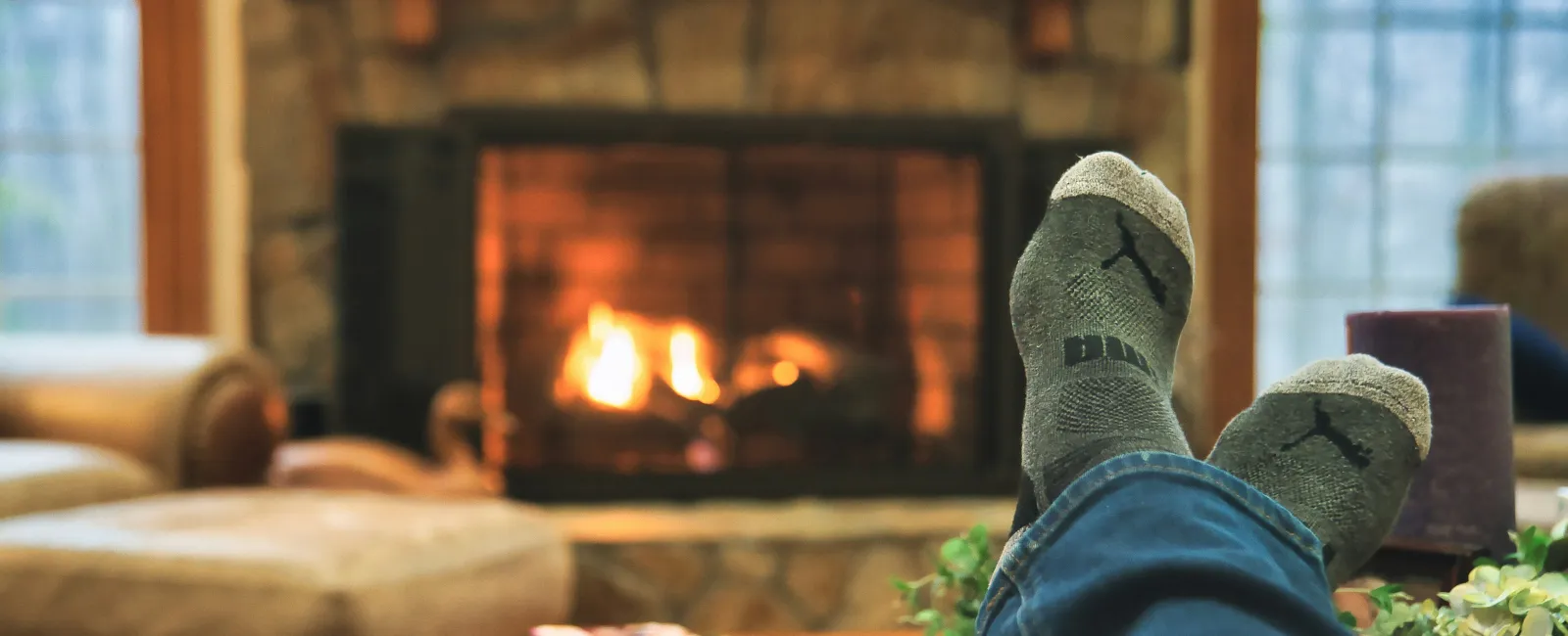 Person relaxing with feet up near a cozy stone fireplace in a warm, inviting living room setting.
