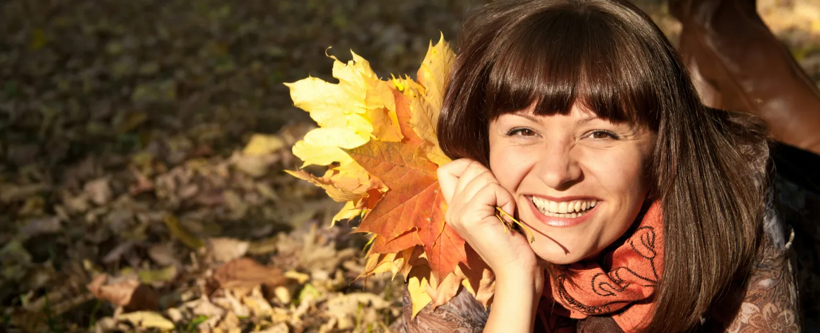 Smiling woman lying on autumn leaves holding a bright yellow and orange maple leaf bouquet outdoors.