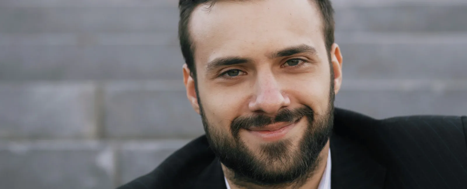 Smiling bearded man in a black blazer sitting outdoors on steps in natural light