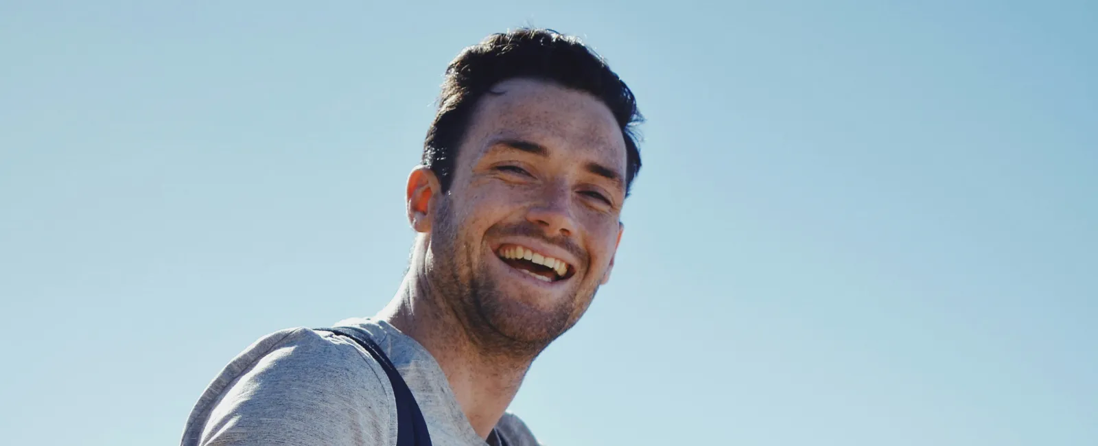 Smiling young man in casual clothes sitting on a rock outdoors under clear blue sky with a backpack.