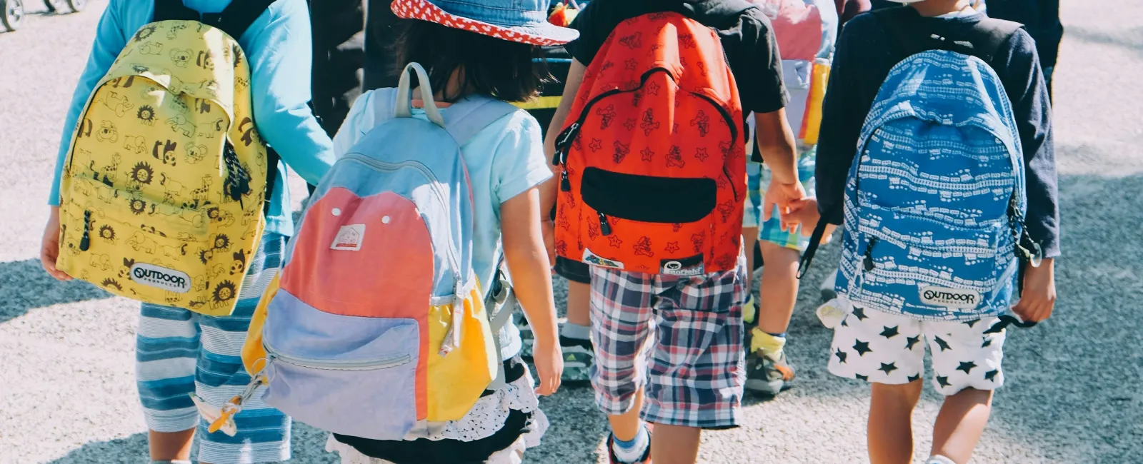 Group of young children wearing colorful backpacks and hats walking outdoors in sunny weather.