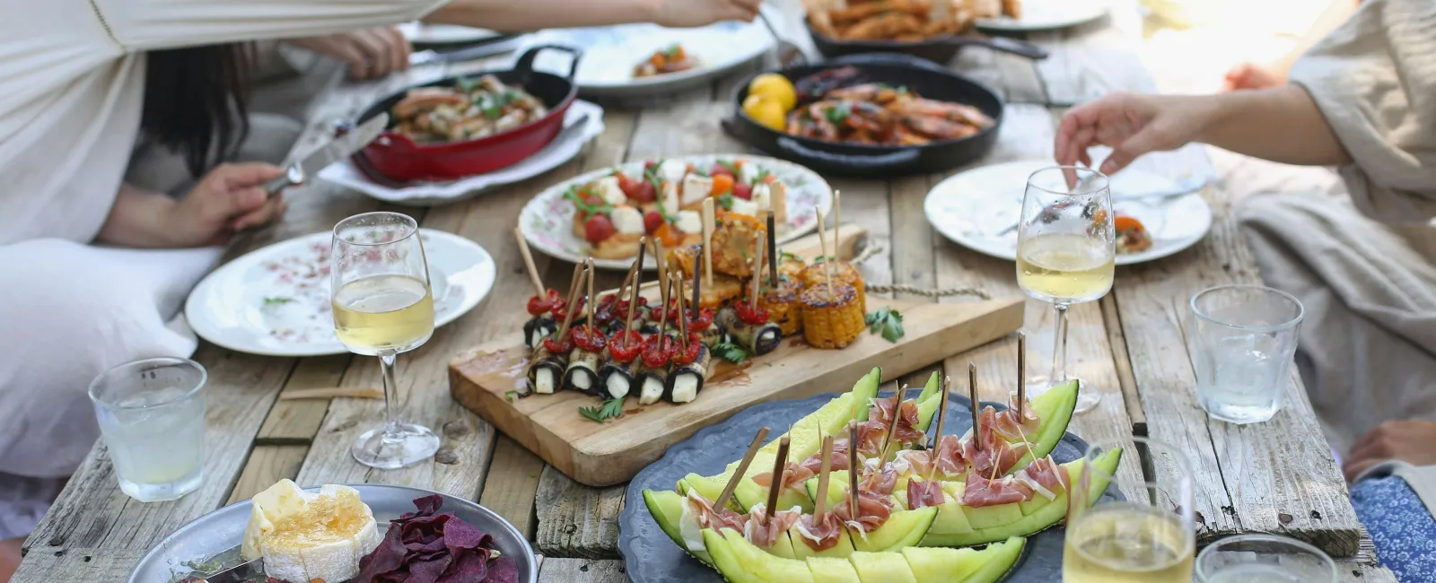 Outdoor wooden picnic table with various appetizers, fruits, nuts, and people enjoying a casual meal together.