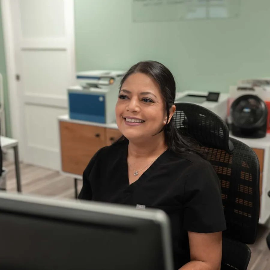 Smiling woman in black scrubs sitting at a desk with computer in a modern office setting.