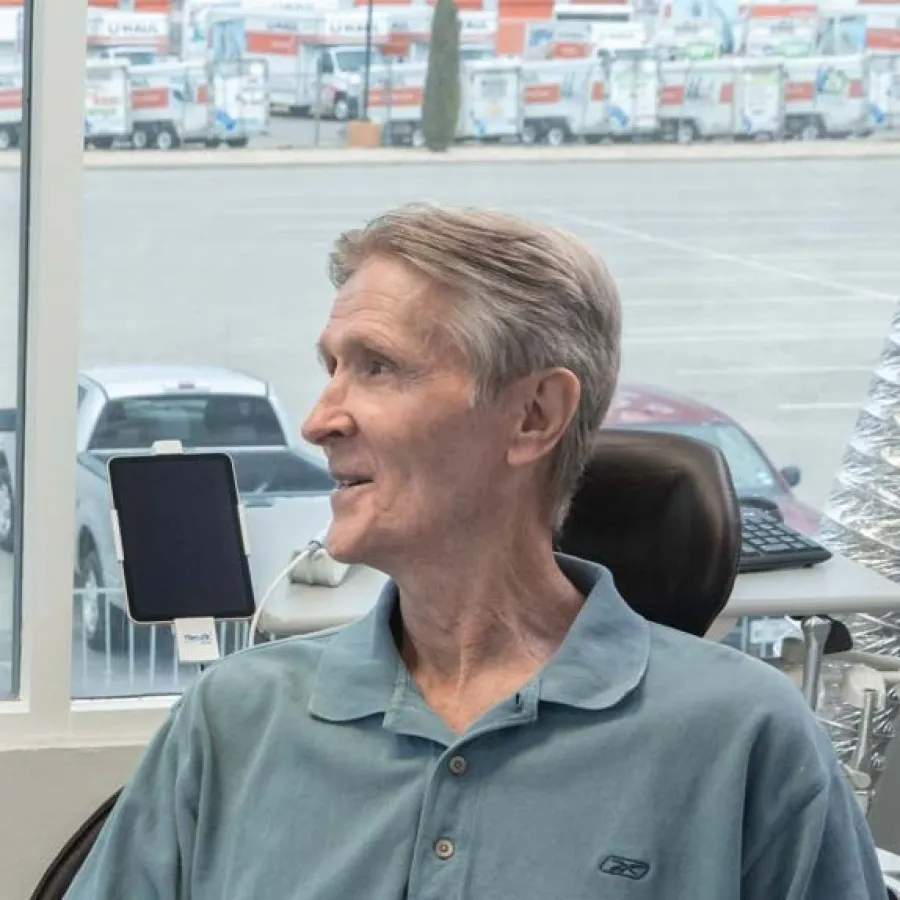Older man in a blue polo shirt sitting indoors near a window with parked trucks visible outside.