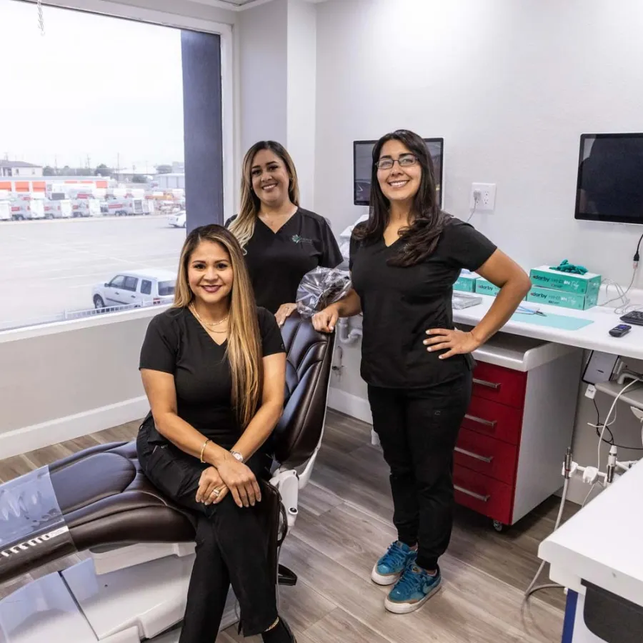 Three female dental professionals in black scrubs smiling inside a modern dental office with equipment.