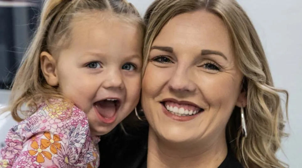 Dr. Hayley Rice holding excited young daughter in floral dress with braided hair indoors.