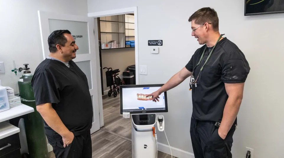 Two dental professionals in black scrubs discuss 3D dental imaging displayed on a screen in a clinic room.