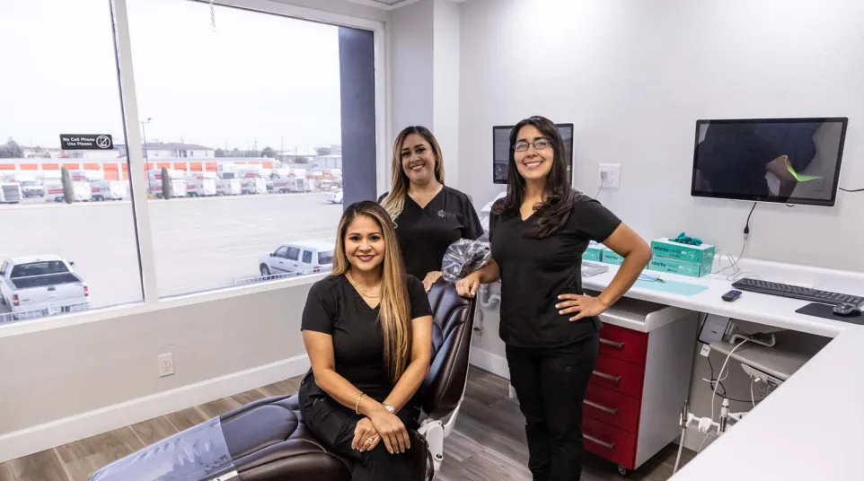 Three smiling female dental professionals posing in a modern dental clinic with a patient chair and computer setup.