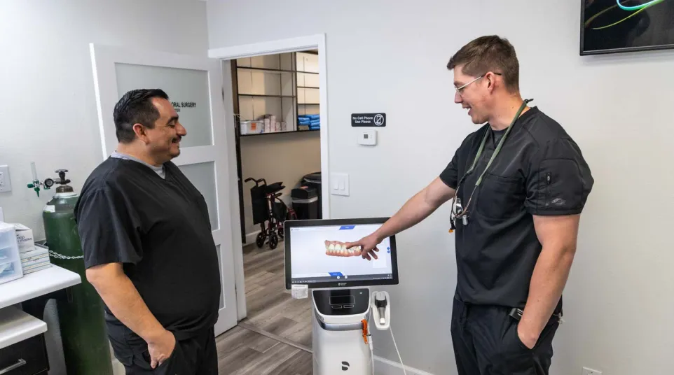 Two dental professionals in black scrubs discuss 3D dental imaging displayed on a screen in a clinic room.