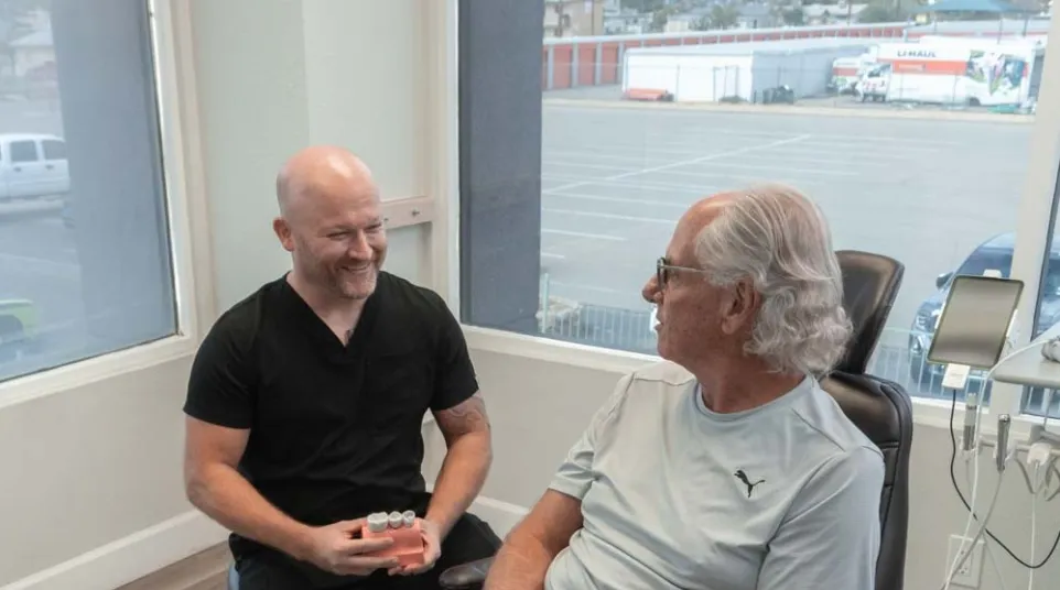 Dentist consulting with male patient sitting in dental chair in a bright clinic with large windows.