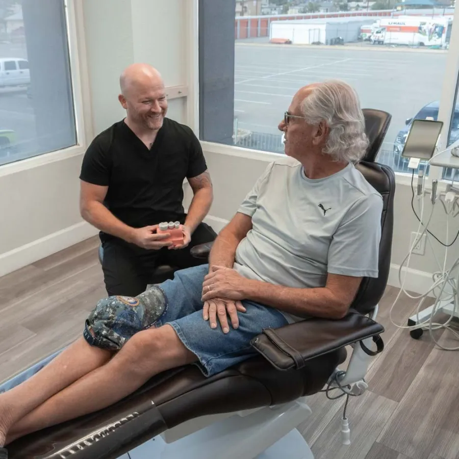 Dentist consulting with male patient sitting in dental chair in a bright clinic with large windows.