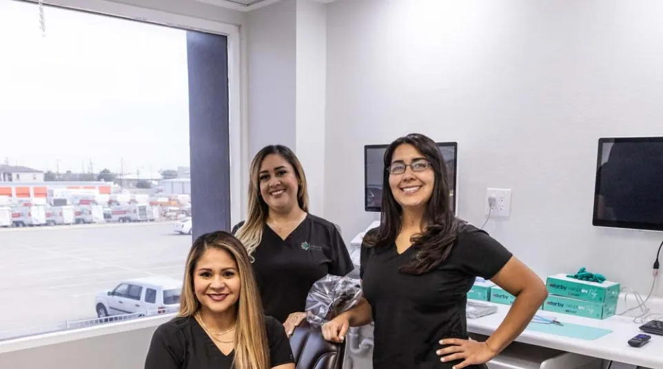 Three female dental professionals in black scrubs smiling inside a modern dental office with equipment.