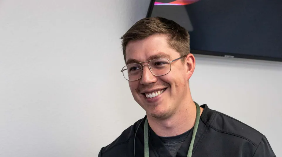 Smiling young man with glasses wearing a black jacket indoors against a white wall and partial TV screen.