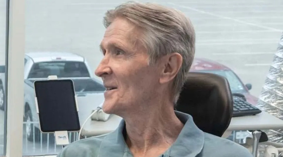 Older man in a blue polo shirt sitting indoors near a window with parked trucks visible outside.