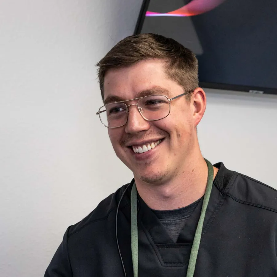 Smiling young man with glasses wearing a black jacket indoors against a white wall and partial TV screen.