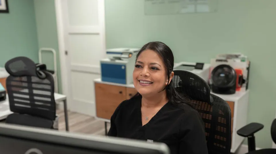 Smiling woman in black scrubs sitting at a desk with computer in a modern office setting.