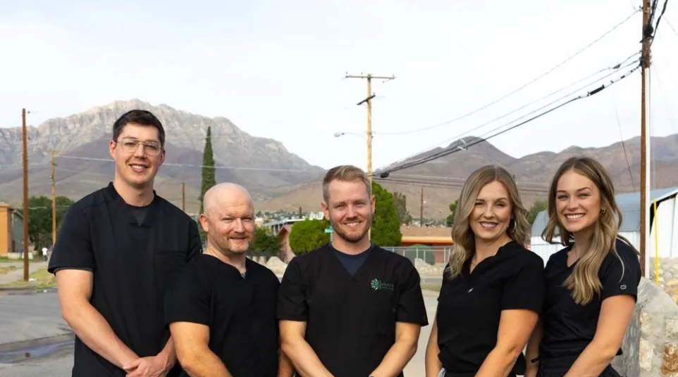 Five healthcare professionals in black scrubs standing outdoors with mountains in the background, smiling at camera.