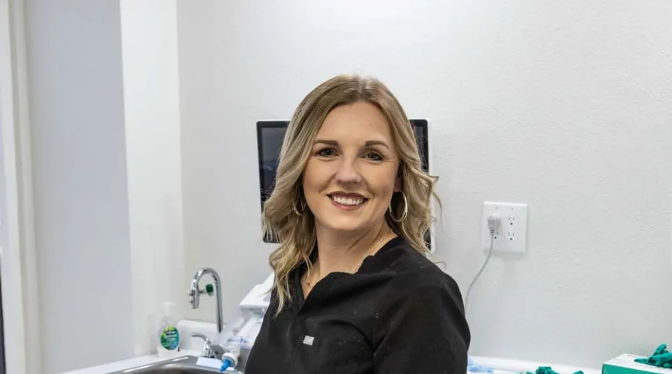 Smiling female dental professional in black scrubs stands next to dental chair in a modern clinic.