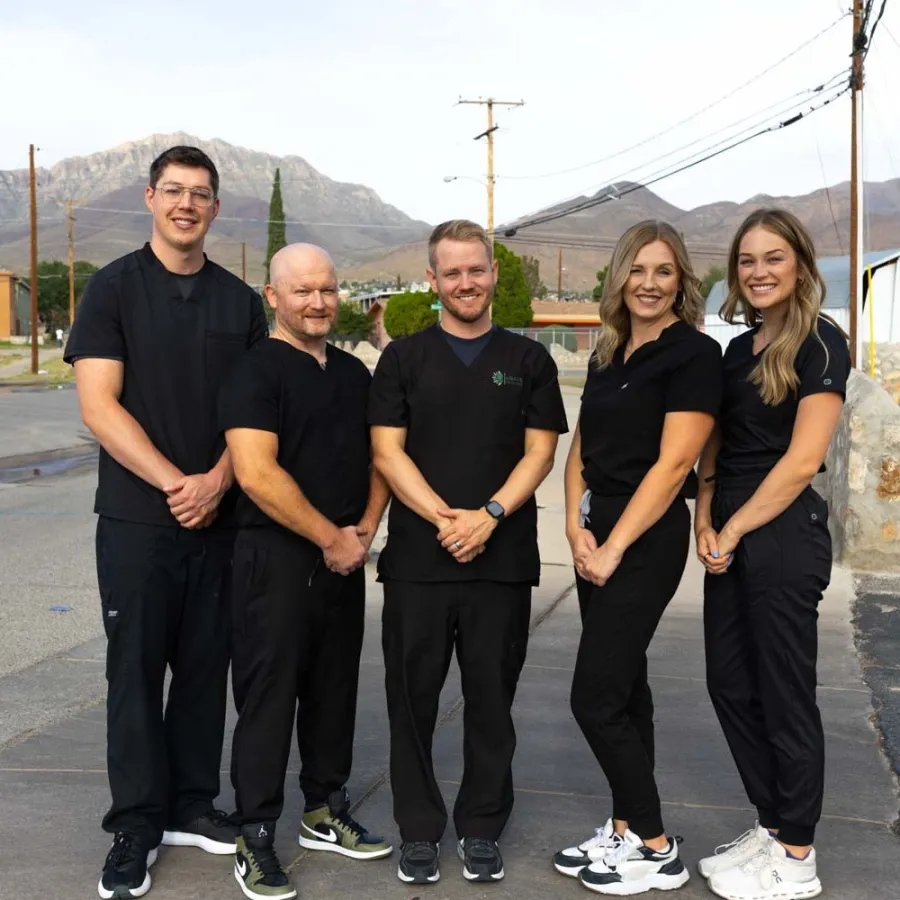 Five healthcare professionals in black scrubs standing outdoors with mountains in the background, smiling at camera.