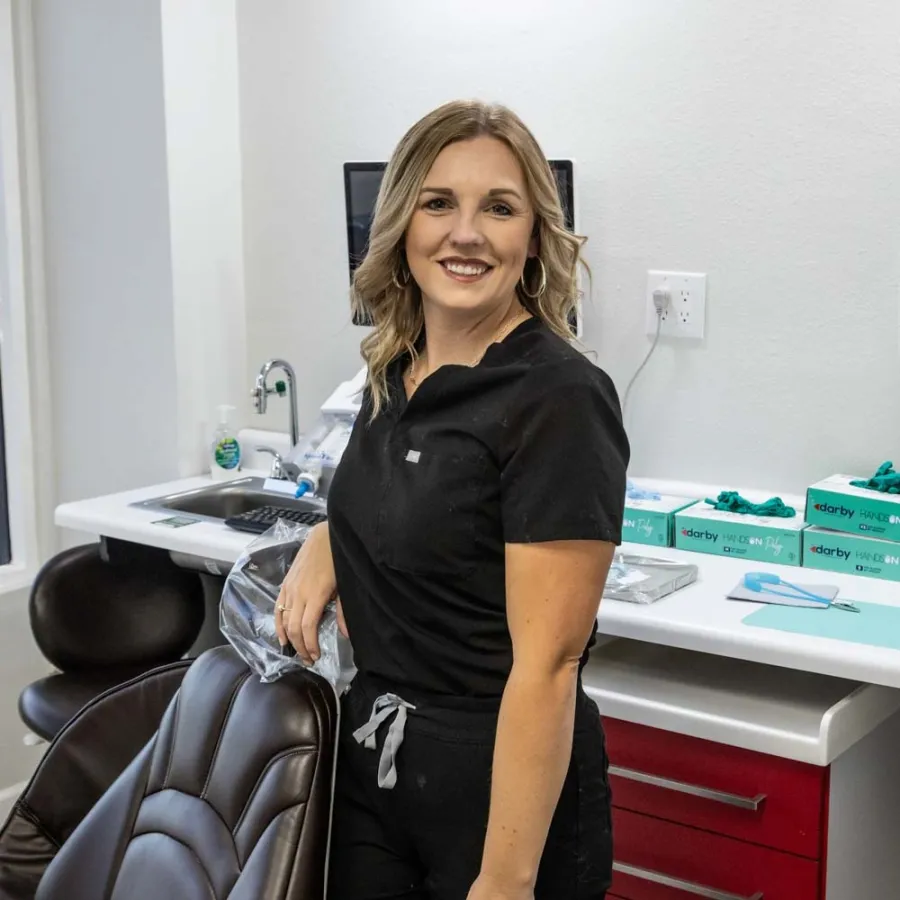 Smiling female dental professional in black scrubs stands next to dental chair in a modern clinic.